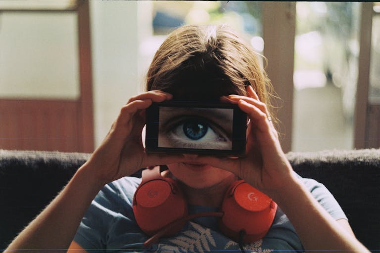 A person with headphones playfully holds a smartphone showing a large eye over their face.