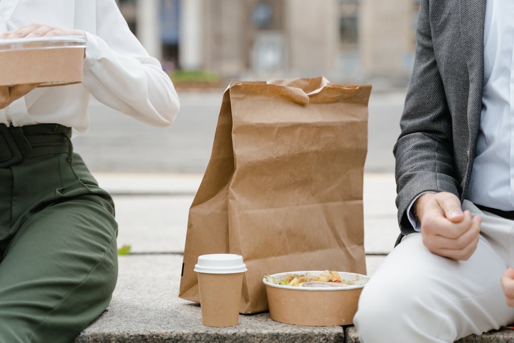 Two people enjoying a casual outdoor lunch with takeaway food packaging and a coffee cup.